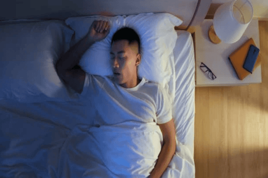 Person sleeping peacefully in bed at night, wearing a white T-shirt, with a bedside table holding glasses, a lamp, and a notebook.