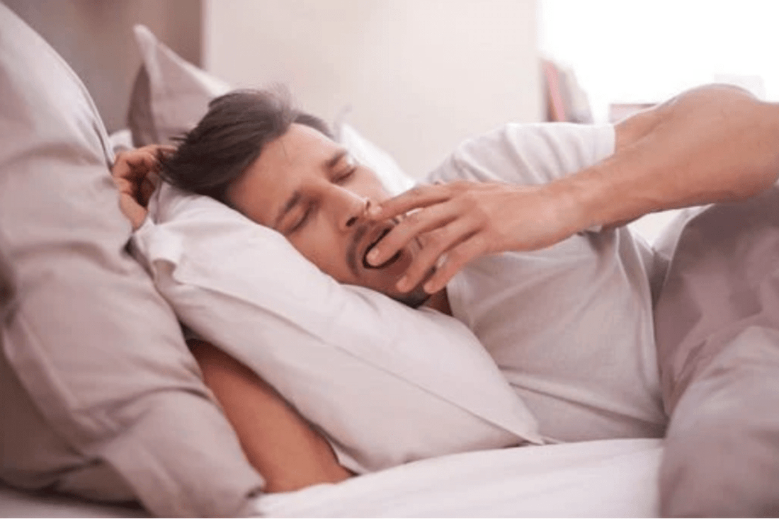 Man waking up in bed with dry mouth, appearing tired and uncomfortable, touching his lips while lying on a pillow.