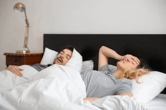 Man snoring loudly in bed while his partner lies awake, covering her face in frustration.