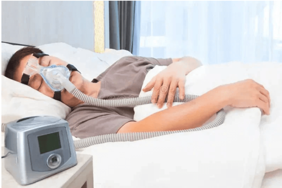 A man is sleeping on his side wearing a full-face CPAP mask connected to a hose and machine on the nightstand, used for treating sleep apnea