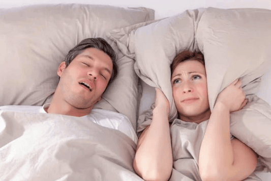 A man is sleeping with his mouth open while snoring, and a frustrated woman beside him is covering her ears with a pillow to block out the noise