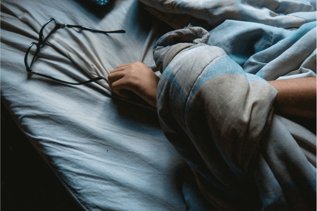 A person is lying in bed under a blue and gray blanket, with one hand visible next to a pair of eyeglasses resting on the bedsheet, suggesting they fell asleep while reading