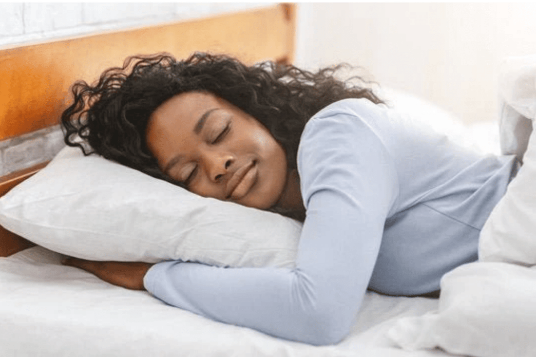 A woman is peacefully sleeping on her side with a gentle smile, resting her head on a white pillow in a cozy, well-lit bedroom
